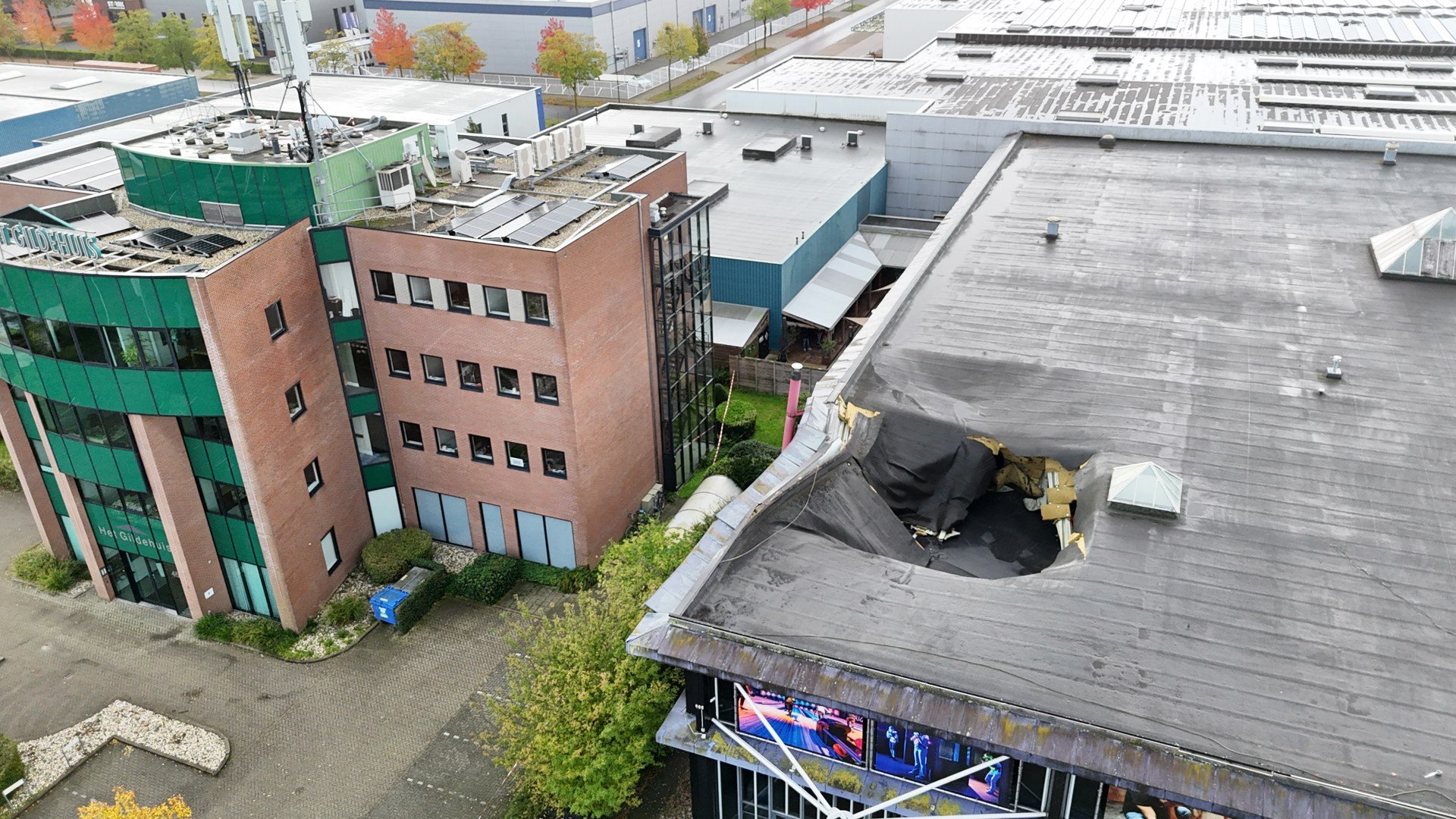 Aerial view of a modern office complex with a damaged flat roof showing a large hole and exposed interior debris on the right side