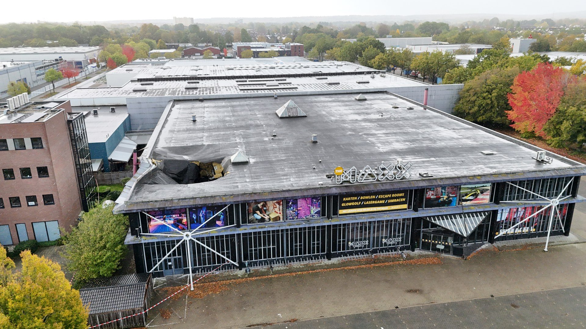 Aerial view of a large building with a damaged flat roof and boarded storefronts along the ground level, autumn trees nearby.