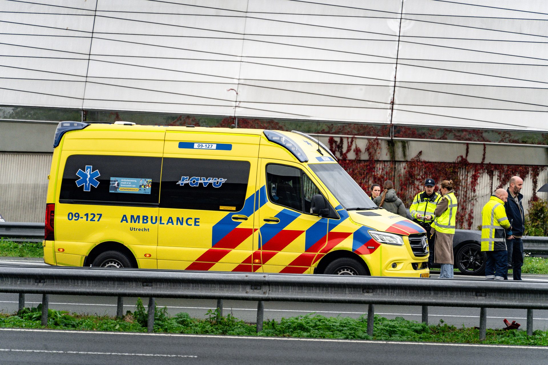 Yellow Dutch ambulance on a highway with responders in neon vests gathered nearby, bus and rail guard in foreground, Utrecht marked on the vehicle.