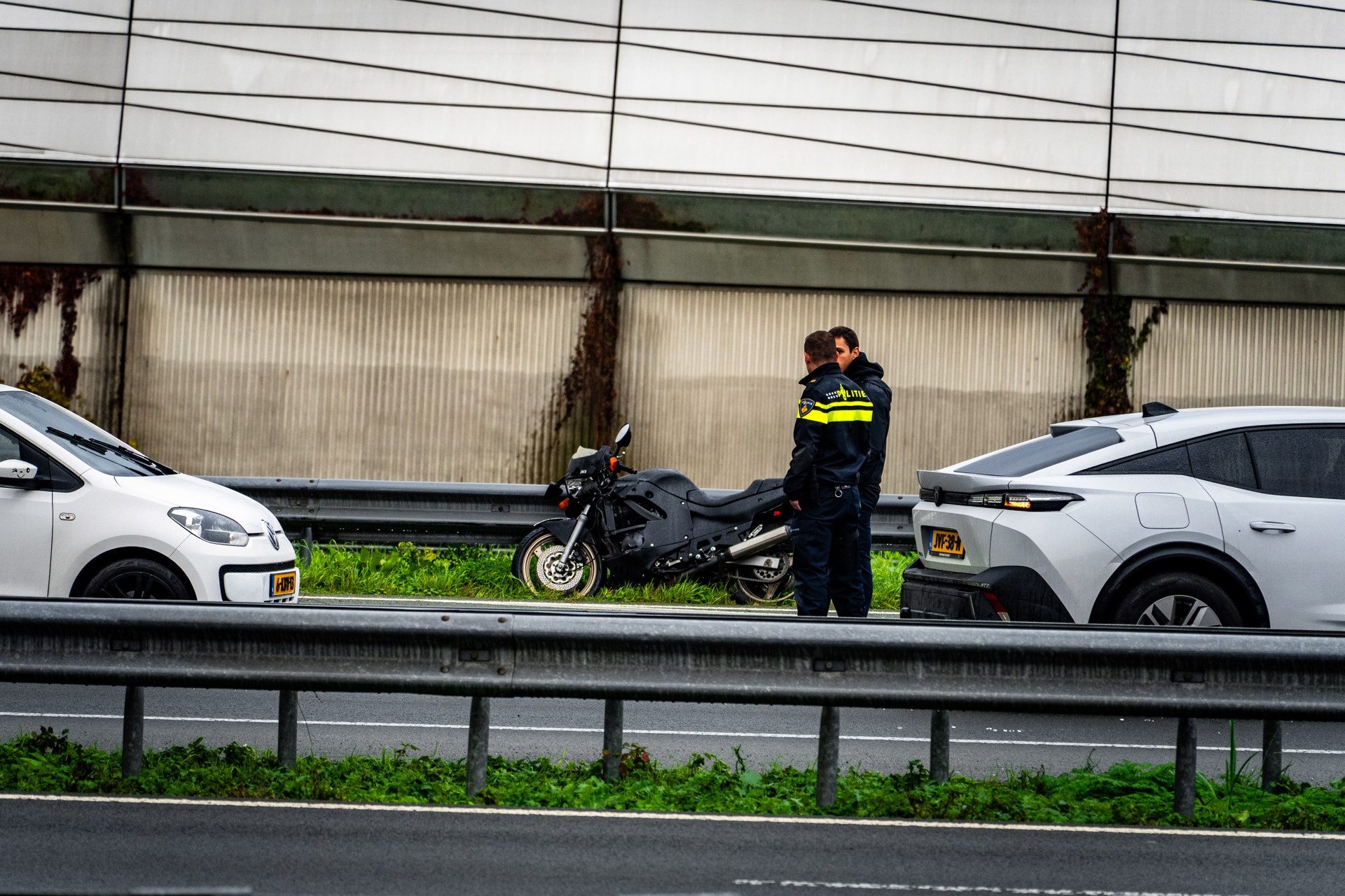 Two police officers in dark uniforms stand beside a black motorcycle on a highway shoulder, near a white car and a gray SUV under a concrete wall fence.