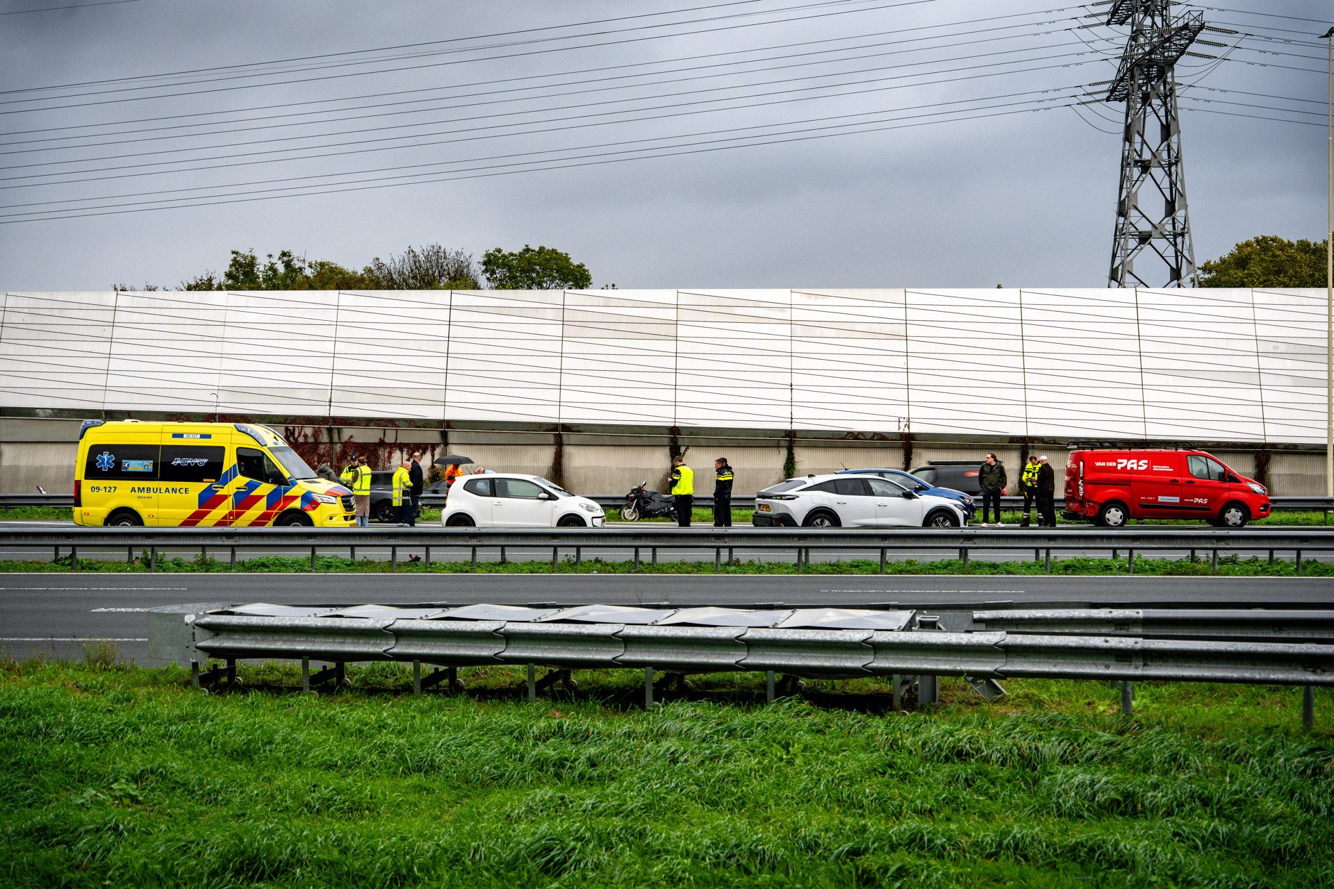 Emergency responders and vehicles along a highway: yellow ambulance, red van, and bystanders on the roadside under a cloudy sky.