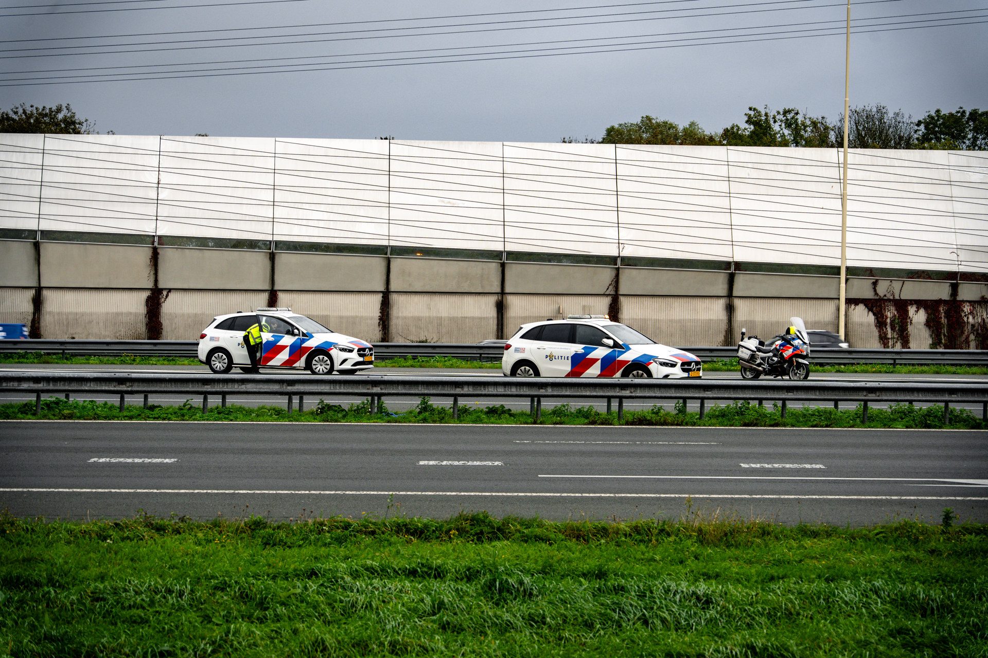 Two Dutch police cars and a police motorcycle parked on a highway beside a tall sound barrier under a gray sky.