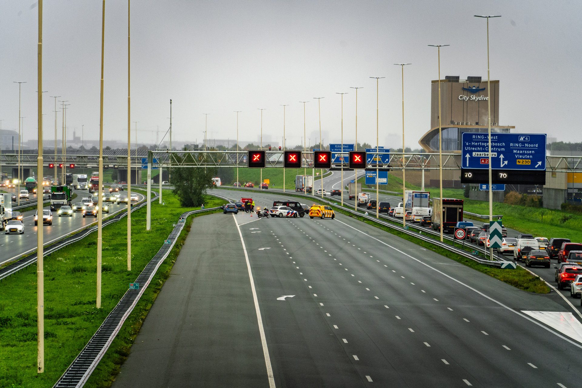 Wide highway scene with multiple lanes and overhead red X signals; traffic on both sides and a large building with 'City Skydive' in the background.