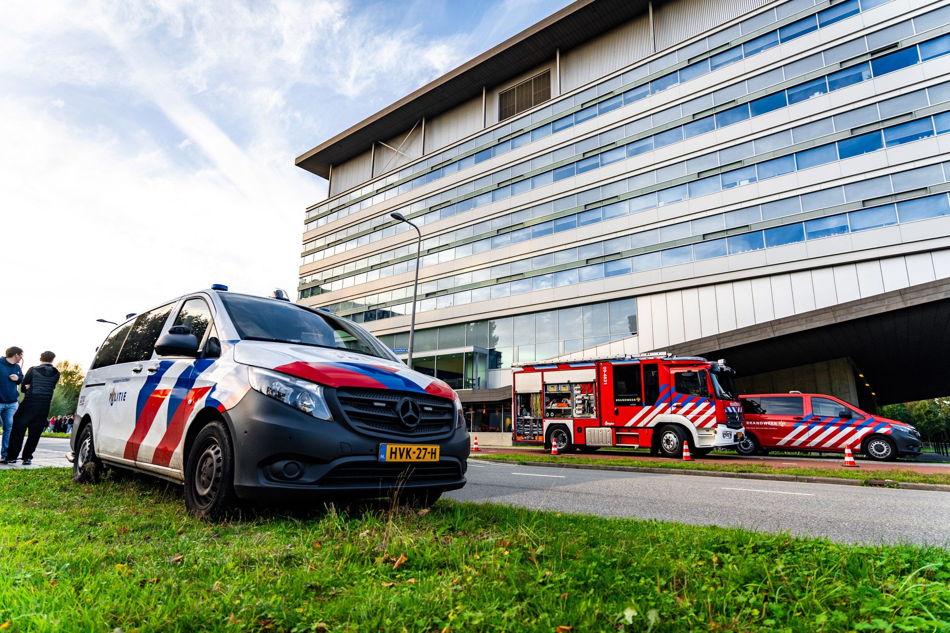Emergency vehicles—police van and fire trucks—parked outside a modern glass office building on a sunny day.