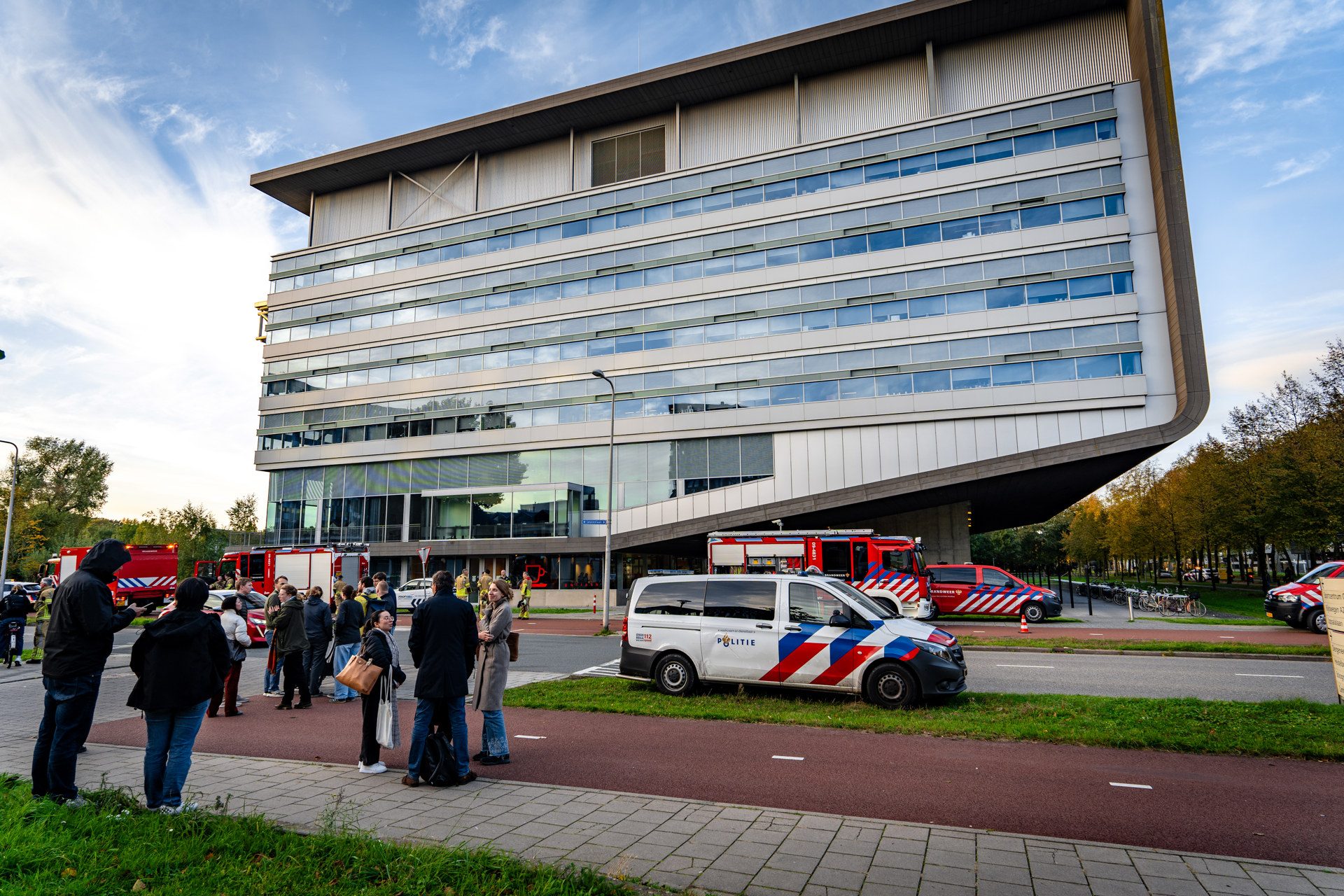 Crowd gathered outside a modern multi-story building with police and fire vehicles parked along the curb, suggesting a public safety drill or event.