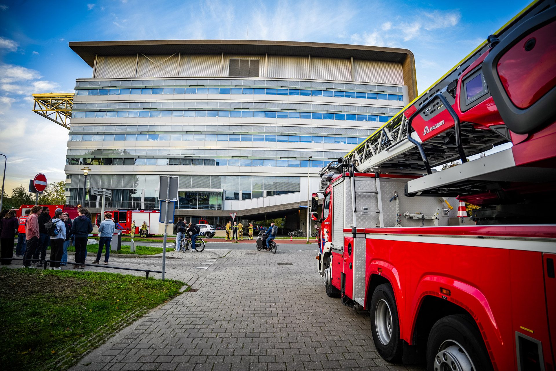 Firefighters and bystanders gather outside a modern office building with a red fire truck and ladder parked on the street.