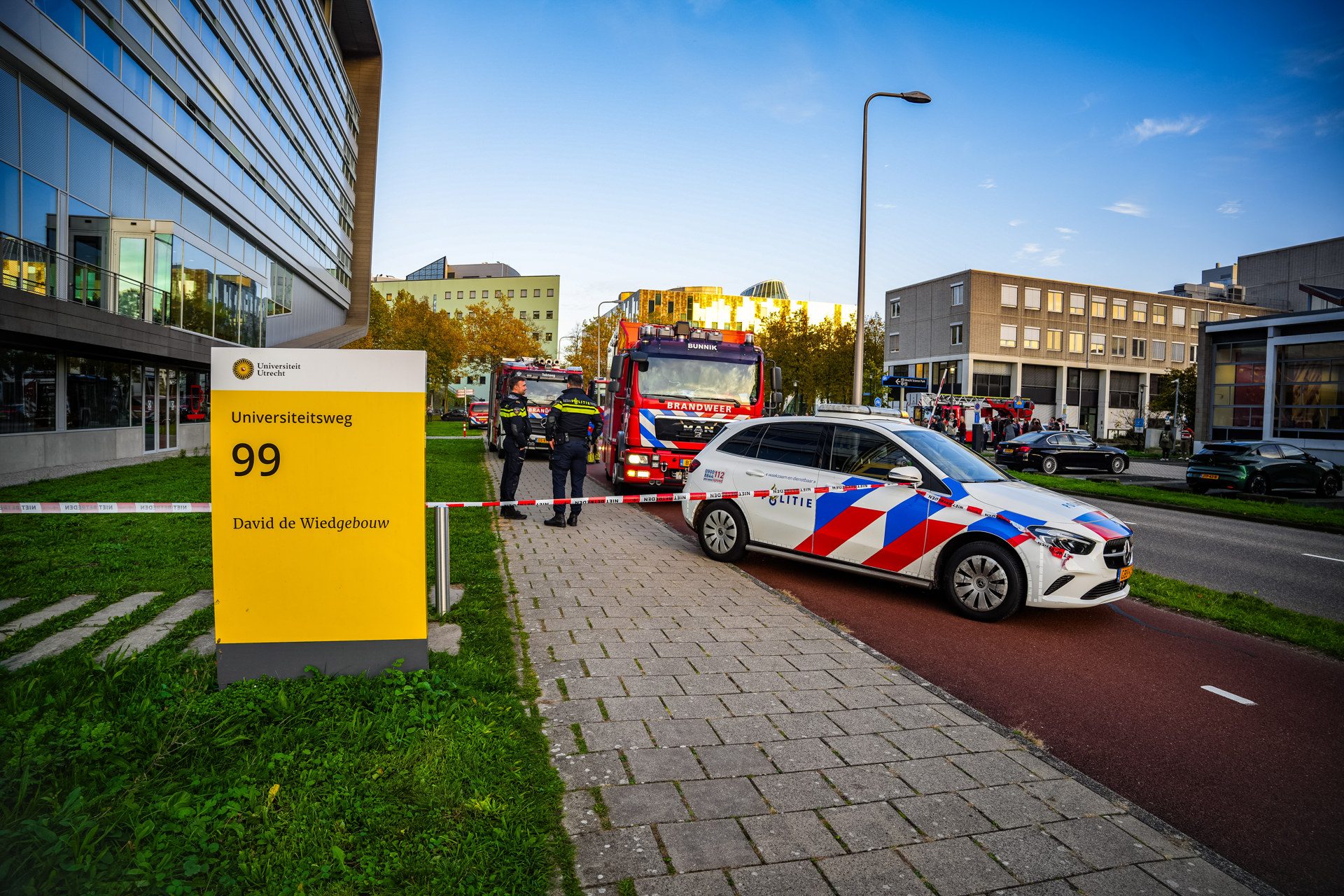 Emergency scene outside a university with police and fire vehicles behind caution tape near modern buildings.