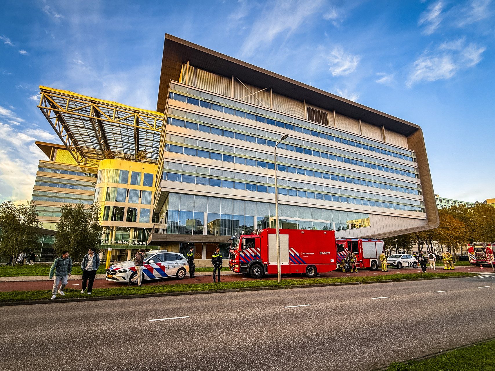 Modern office building with a large glass facade and a yellow-toned overhead structure, under a blue sky, with emergency vehicles nearby.